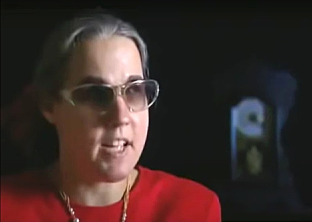 Vicki Umipeg, mulher cega com cabelos grisalhos, usando óculos escuros e blusa vermelha, falando com expressão animada e serena durante depoimento sobre visão espiritual.
